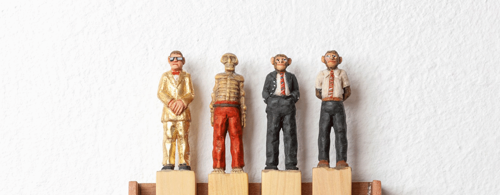 Vier kleine Holzfiguren mit Affengesichtern in unterschiedlichen Anzügen stehen auf vier Holzklötzen in einem Holzrahmen an einer weißen Wand