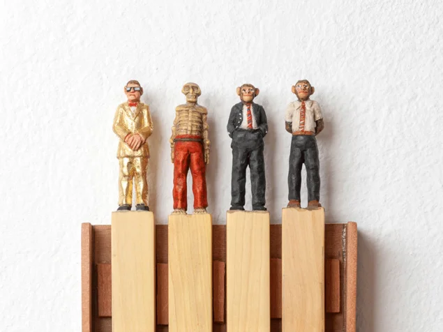 Vier kleine Holzfiguren mit Affengesichtern in unterschiedlichen Anzügen stehen auf vier Holzklötzen in einem Holzrahmen an einer weißen Wand