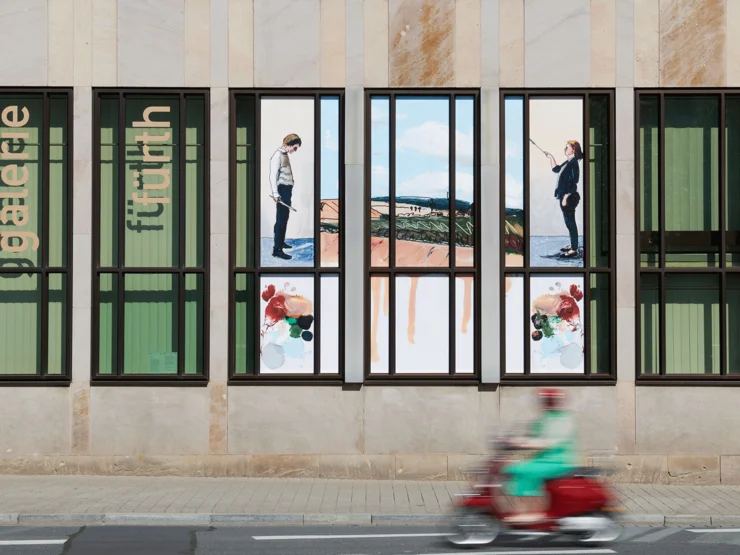 Fassade der kunst galerie fürth mit großen Fenstern, in denen Bilder von zwei Personen und Landschaften zu sehen sind, davor unscharfe Person auf rotem Motorroller