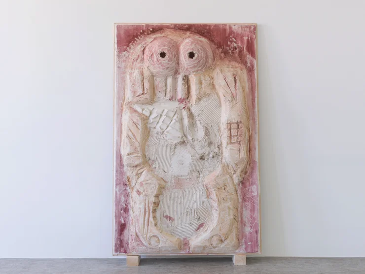 Relief mit abstrakter, symmetrischer Figur mit zwei großen Augen auf einem rechteckigen, rosa-weißen Hintergrund