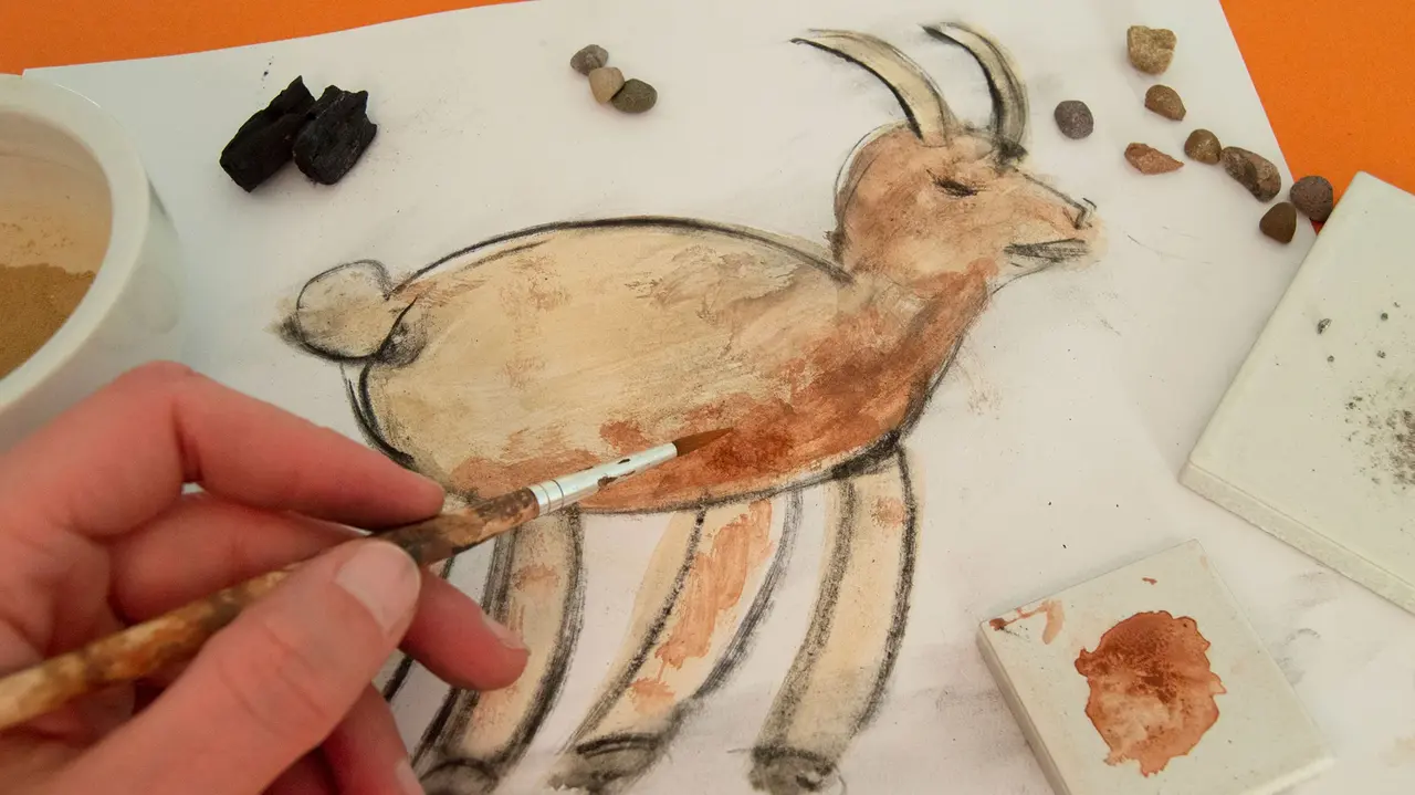 Eine Hand mit Pinsel malt eine Ziege. Auf dem Papier liegen verschiedene Steine.