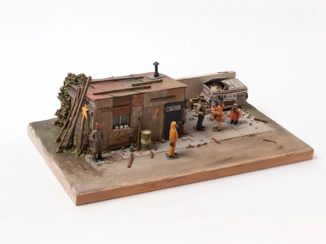 Miniaturszene mit einem kleinen, verwitterten Holzhaus, mehreren Figuren in Winterkleidung und einem alten Auto auf einem rechteckigen Sockel.