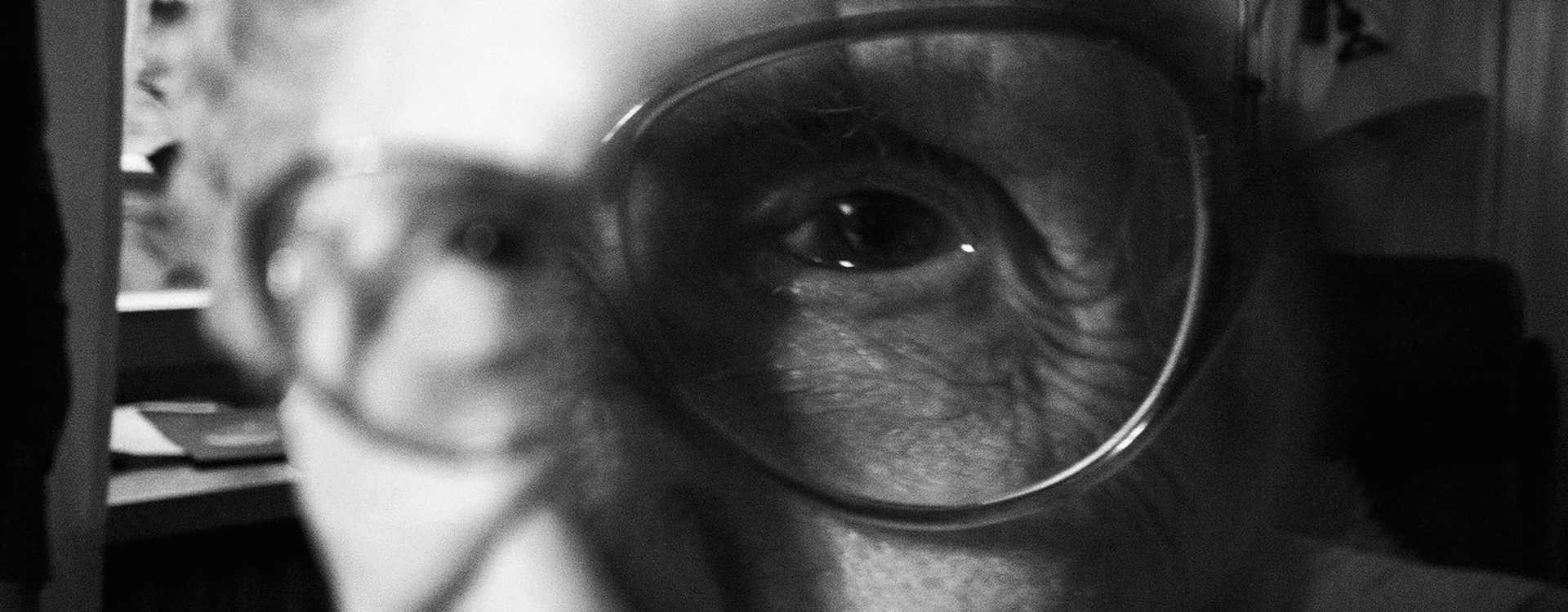 Ältere Frau mit Brille sitzt an einem Tisch in einem Raum mit Fenster und Vorhang. 
