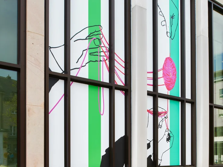 Fensterfront der Kunstgalerie mit moderner Kunst, die Hände zeigt, die pinken Faden halten, vor weißem Hintergrund mit grünen senkrechten Streifen