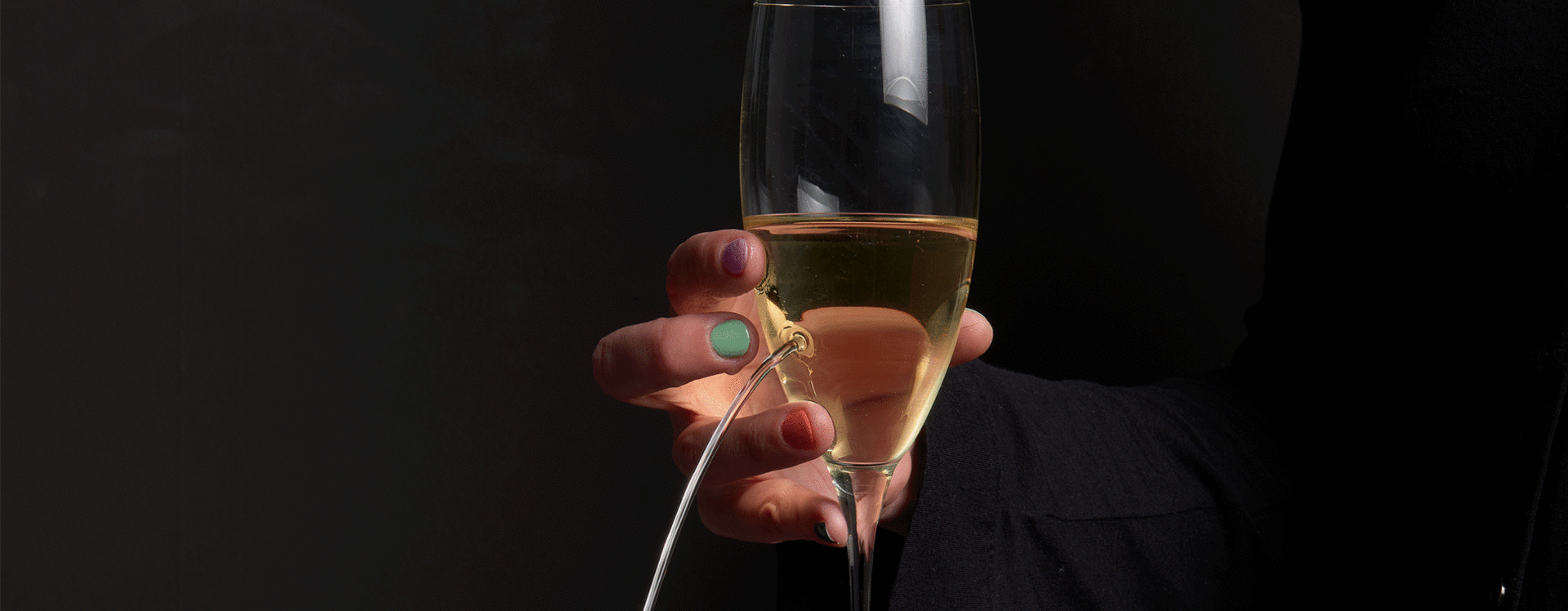 Hand mit bunten Fingernägeln hält ein Sektglas, aus dem Flüssigkeit in einem Bogen herausfließt.