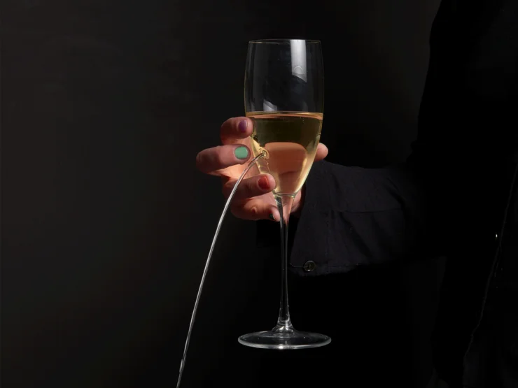 Hand mit bunten Fingernägeln hält ein Sektglas, aus dem Flüssigkeit in einem Bogen herausfließt.